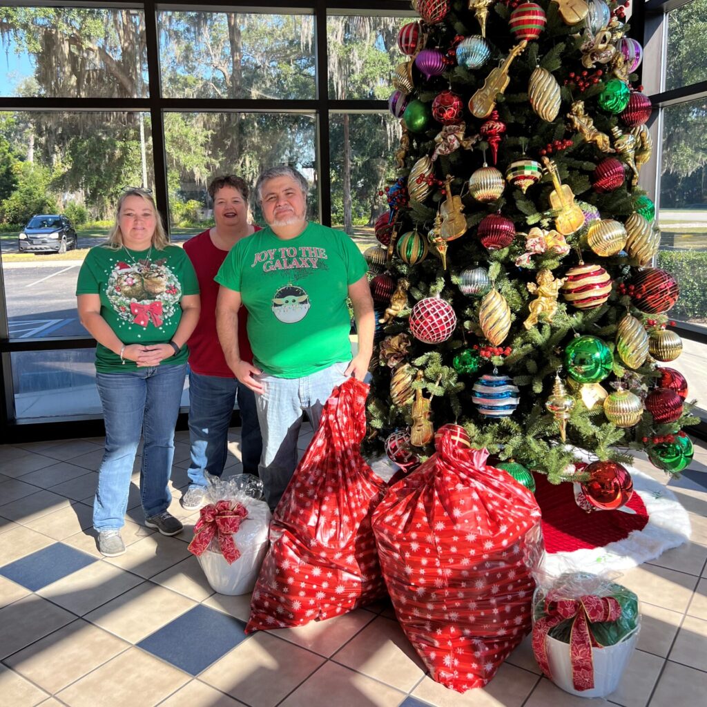 Group of young people standing next to tall Christmas tree with bags of presents in foreground