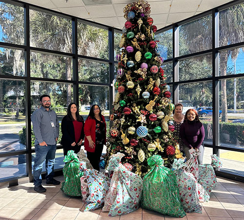 Employees stand next to large Christmas tree with large bags of gifts in front of them